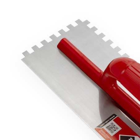 Une truelle crantée avec un manche rouge repose sur une surface blanche. Il comporte une lame en métal avec des encoches carrées