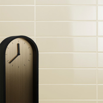 Un reloj minimalista con marco negro no muestra números, solo marcas de hora negras, sobre un fondo de pared de azulejos beige.
