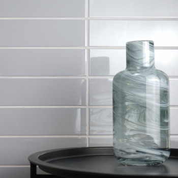 Eine gewirbelte Glasflasche ruht auf einem schwarzen Tablett vor einer glänzend weißen U-Bahn-Fliesenwand und reflektiert das Um