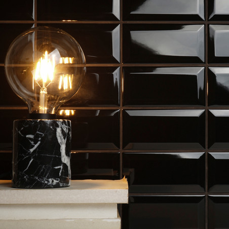 Uma lâmpada brilhante fica sobre uma base de mármore preto, iluminando um fundo de parede de azulejos pretos brilhantes que refl