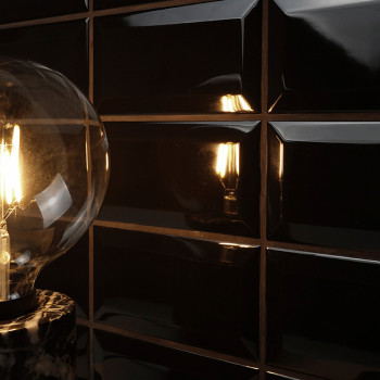 Uma lâmpada incandescente emite luz quente, refletida em ladrilhos retangulares pretos brilhantes. A configuração cria um ambien