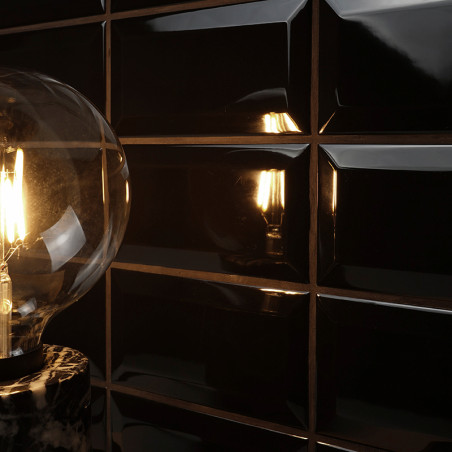 Uma lâmpada incandescente emite luz quente, refletida em ladrilhos retangulares pretos brilhantes. A configuração cria um ambien