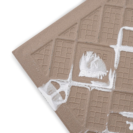 A textured brown ceramic tile is etched with grid patterns and white paint splatters, featuring the text `MADE IN SPAIN.`