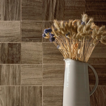 Una jarra de cerámica blanca contiene flores secas de color beige y violeta, colocadas contra una pared de azulejos marrones con