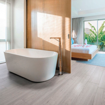 Eine weiße freistehende Badewanne steht auf hellem Holzboden neben einer Holztrennwand und bietet Blick auf ein Schlafzimmer mit