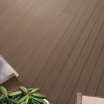 Des carreaux marron avec des rainures parallèles se trouvent à l'extérieur, à côté d'une chaise en osier et d'une plante verte, 