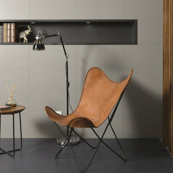Op een donkere tegelvloer staat een leren vlinderstoel, naast een moderne staande lamp en een kleine ronde tafel. Tegen een bete
