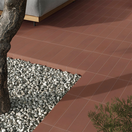 Los azulejos de terracota forman una superficie de patio limpia adyacente a un área de grava con un tronco de árbol. Un sofá gri