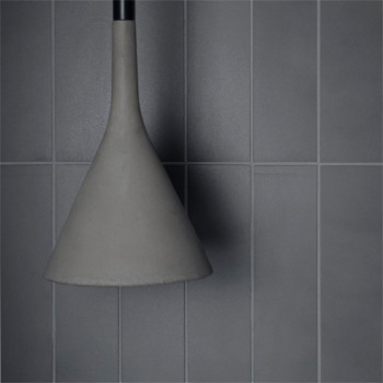 Una lámpara en forma de cono cuelga sobre un fondo de azulejos grises rectangulares y lisos, creando una estética industrial min