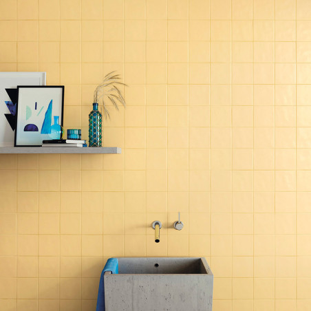 An einer mattgelb gefliesten Wand steht ein minimalistisches Betonwaschbecken mit einem blauen Handtuch unter einem Regal mit Ku
