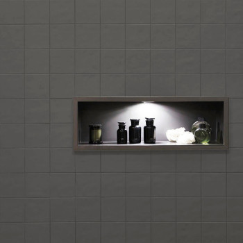 Une étagère encastrée avec des bouteilles noires et un vase vert sous un éclairage tamisé se trouve dans un mur carrelé gris fon