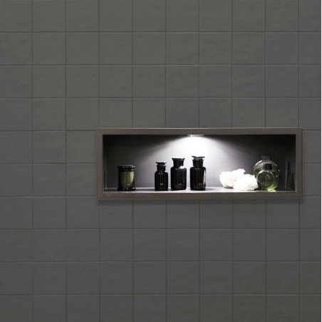 Une étagère encastrée avec des bouteilles noires et un vase vert sous un éclairage tamisé se trouve dans un mur carrelé gris fon