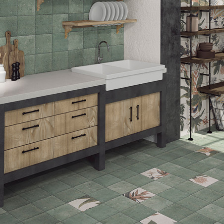 A kitchen counter features wooden drawers and a large sink, surrounded by earthy green tiles with floral patterns, in a rustic s