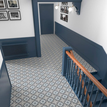 Des carreaux géométriques à motifs recouvrent le sol d'un couloir, bordé de lambris bleus et d'un escalier. Des photographies mo