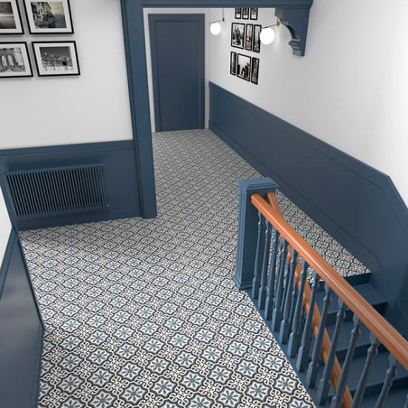 Des carreaux géométriques à motifs recouvrent le sol d'un couloir, bordé de lambris bleus et d'un escalier. Des photographies mo
