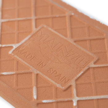 O ladrilho de terracota exibe `MAUNZU` e `MADE IN SPAIN.` O ladrilho apresenta um padrão geométrico de grade com leve desgaste.