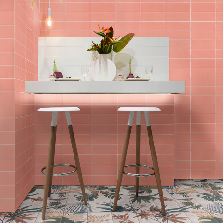 Dois bancos de madeira ficam em um balcão branco adornado com um vaso de flores, contra paredes de azulejos rosa e piso com moti