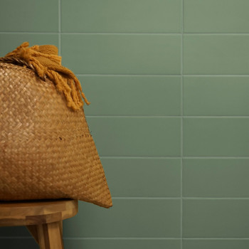Een geweven mand met kwastjes staat op een houten krukje tegen een achtergrond van gladde, groene tegels.