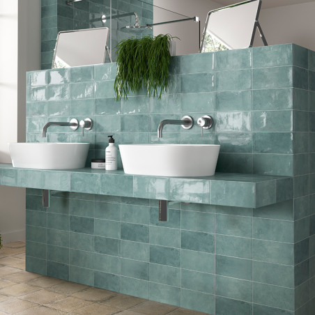 Los lavabos blancos descansan sobre una encimera de azulejos verde azulado, con grifos encima. Espejos y una planta adornan el t