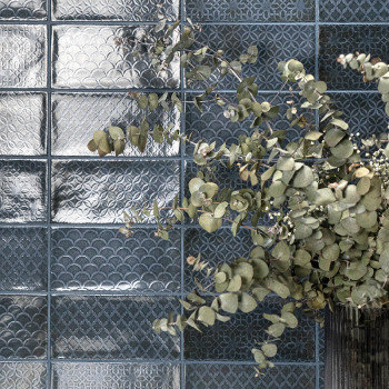 Las ramas de eucalipto verde se extienden sobre azulejos de vidrio con patrones geométricos en relieve, creando un contraste ele