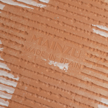 Een oranjebruine keramische tegel, getextureerd met lineaire patronen. De tekst `MAINZU MADE IN SPAIN` is in het oppervlak geste