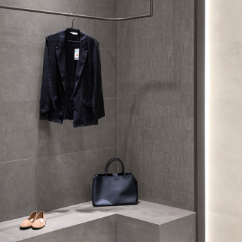 A black jacket hangs on a hook beside a black handbag on a gray tiled bench, with beige shoes on the floor below.