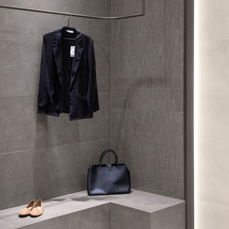 A black jacket hangs on a hook beside a black handbag on a gray tiled bench, with beige shoes on the floor below.