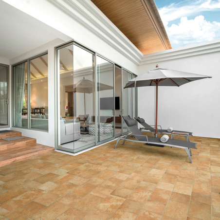 Deux chaises longues avec serviettes reposent sous un grand parasol sur un patio carrelé beige, adjacent à une pièce moderne aux