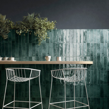 Groene, glanzende tegels vormen een muurdecor onder hangplanten. Witte metalen barkrukken rusten tegen een houten aanrecht, waar