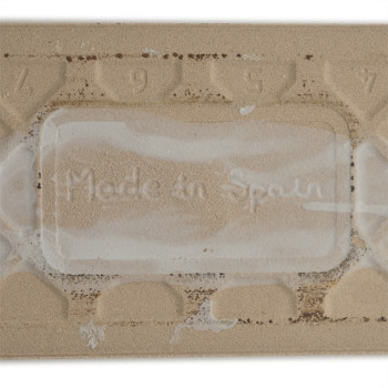 Op een rechthoekige beige tegel staat de tekst `Made in Spain` in reliëf, omringd door een structuurpatroon met de cijfers 9, 5,