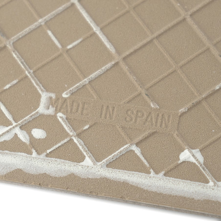 Un carreau beige présente un motif de grille géométrique avec quelques résidus blancs. Le texte indique « FABRIQUÉ EN ESPAGNE ».