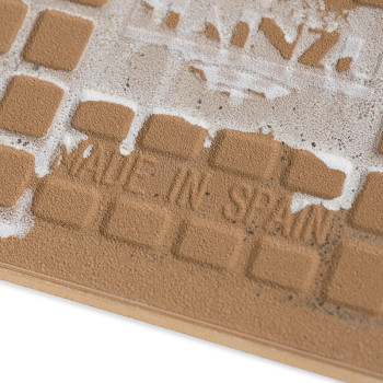 Un azulejo de cerámica marrón tiene cuadrados texturizados que muestran signos de desgaste. El texto impreso dice `MADE IN SPAIN