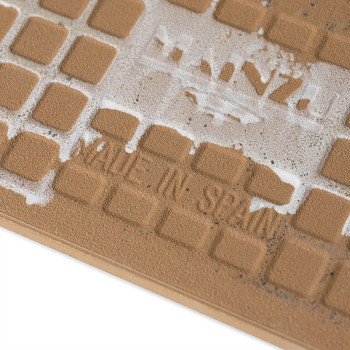 Un azulejo marrón con motivos cuadrados en relieve, ligeramente desgastado, que muestra el texto “MADE IN SPAIN” y logotipos par