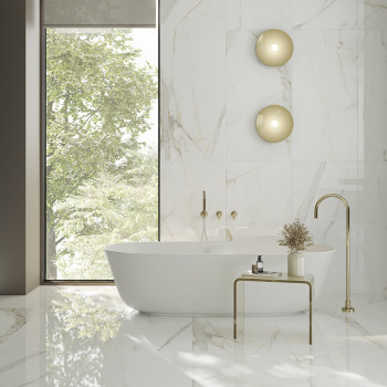 A freestanding white bathtub rests on a glossy, marble-tiled floor, surrounded by natural light from a large window and minimal 