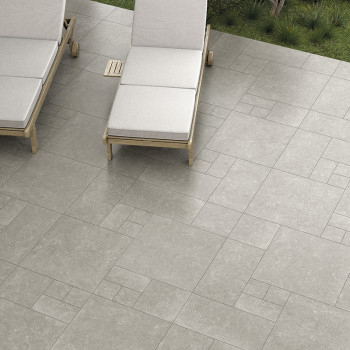 Graue Steinfliesen bilden ein geometrisches Muster auf einer Terrasse, daneben stehen zwei gepolsterte Liegestühle, umgeben von 