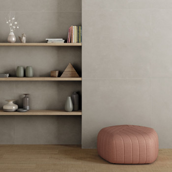 Una otomana rosa descansa sobre un piso de madera junto a estantes empotrados minimalistas que sostienen jarrones, libros y pequ