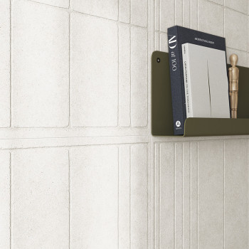 Un azulejo de pared blanco texturizado forma un patrón geométrico. Un estante de color verde oliva contiene libros, incluido `ar