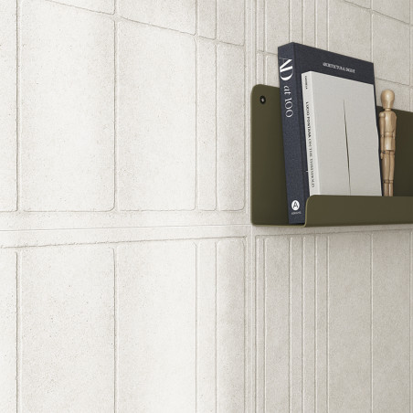 Un azulejo de pared blanco texturizado forma un patrón geométrico. Un estante de color verde oliva contiene libros, incluido `ar