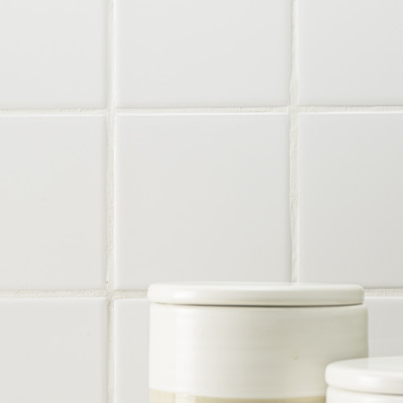 Two white ceramic jars with lids sit against a backdrop of plain white, square tiles with visible grout lines.