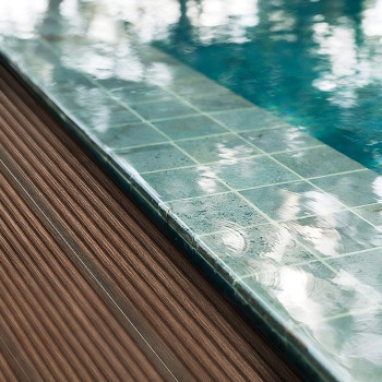 A grid of turquoise stone tiles borders a reflective swimming pool beside a textured wooden deck.