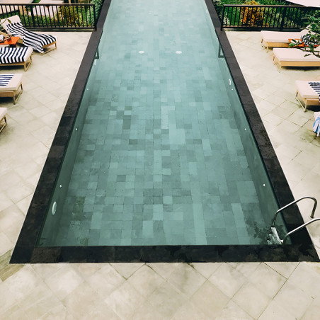 Rectangular stone tiles form a pool bottom beneath clear water, surrounded by lounge chairs and vibrant green plants.