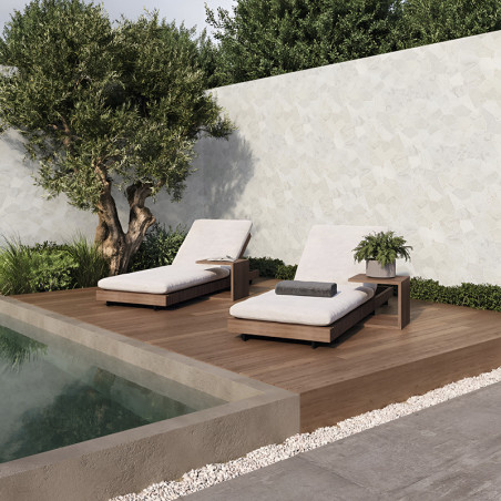 Two cushioned lounge chairs rest on a wooden deck beside a pool, surrounded by lush greenery and a stone wall.