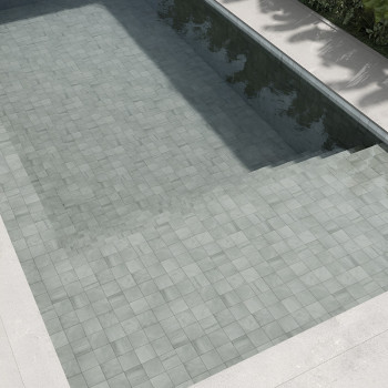 Light gray stone tiles form a submerged pool floor, reflecting sunlight, surrounded by concrete edges and green foliage.