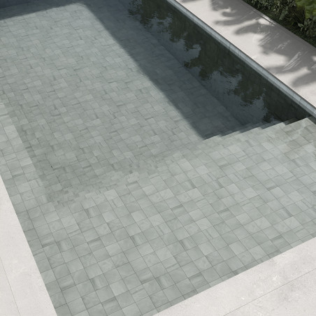 Light gray stone tiles form a submerged pool floor, reflecting sunlight, surrounded by concrete edges and green foliage.
