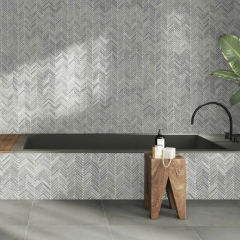 Gray stone tiles form a herringbone pattern wall above a bathtub, complemented by wood stools and a plant.