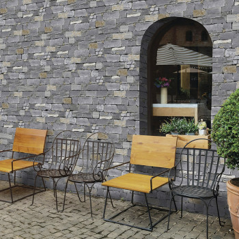 Stone tiles form a rustic wall, framing a large arched window, bordered by iron chairs and potted plants on a cobblestone path.