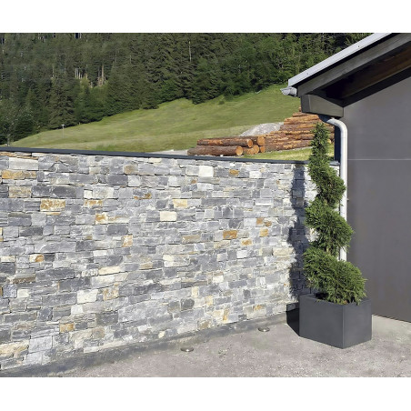 Stone wall standing amidst a yard, bordered by lush grass and trees, adjacent to a modern building corner.