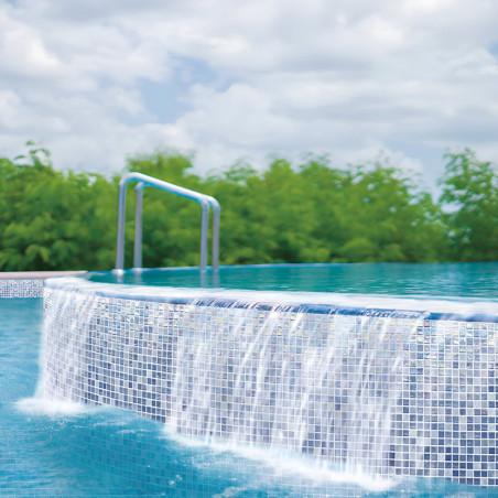 Water cascades over a glass mosaic infinity pool edge, set against lush greenery and a cloudy sky.
