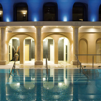 Pool mit reflektierenden Bögen und Säulen. Warme Lichter beleuchten die Gebäudefassade in einer ruhigen Indoor-Spa-Umgebung.
