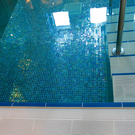 Ein Glasmosaik-Pool reflektiert das Sonnenlicht und erzeugt schimmernde Muster in einer Schwimmhalle mit sichtbaren Metallgeländ